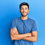 young handsome man wearing casual tshirt over blue background happy face smiling with crossed arms looking at the camera. positive person.